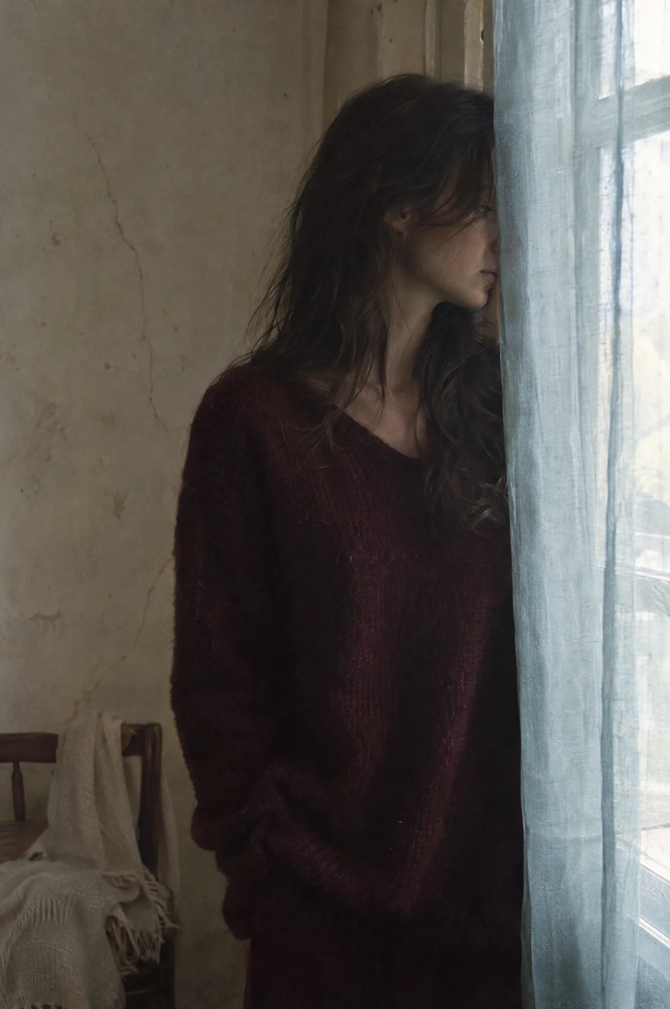 A woman in a wine-coloured knit standing half in shadow beside a tall window, her face turned away behind a sheer blue-grey curtain.
